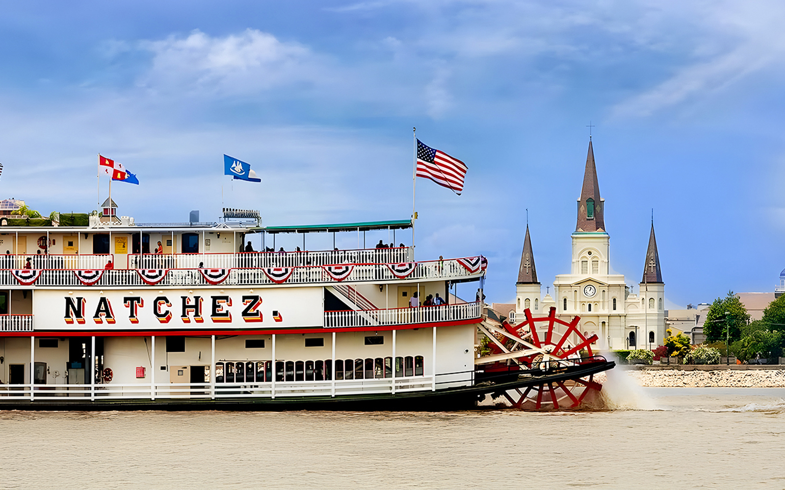 Steamboat Natchez cruising on New Orleans waters with city skyline in the background.