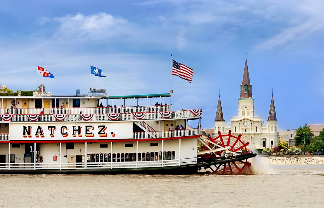 Steamboat Natchez cruising on New Orleans waters with city skyline in the background.