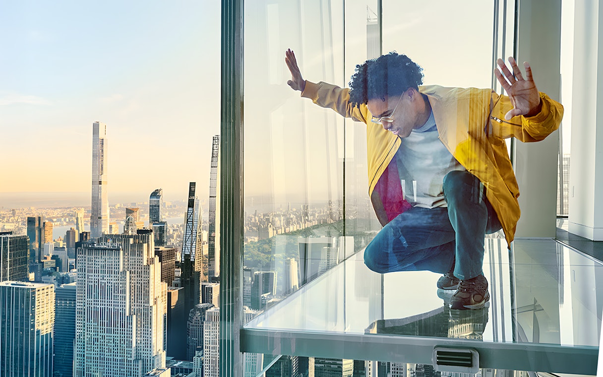 Man crouching on glass floor with view of New York City skyline at SUMMIT OV.