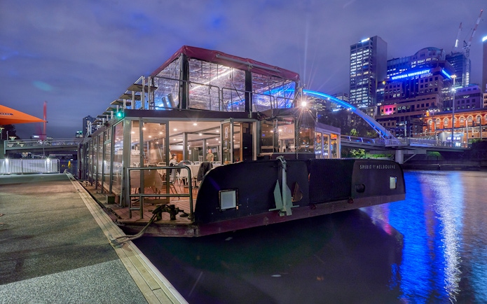 Dinner cruise boat docked on Yarra River, Melbourne, with city skyline in the background.