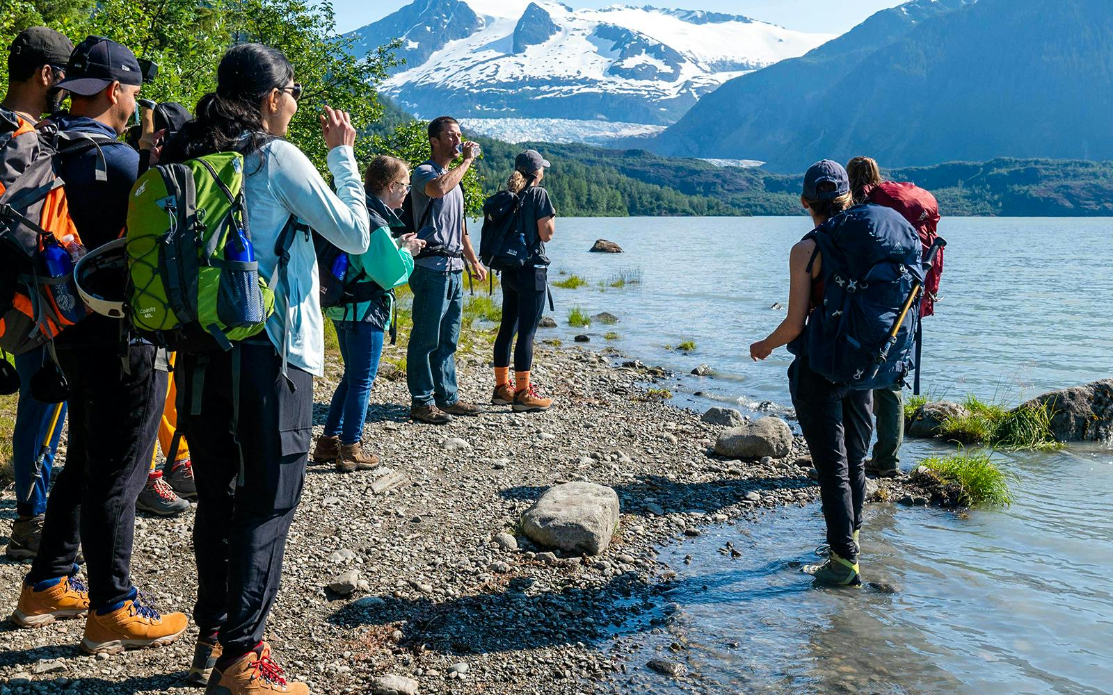Group hiking along Mendenhall Glacier lake with snowy mountains in the background.