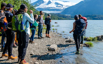 Group hiking along Mendenhall Glacier lake with snowy mountains in the background.