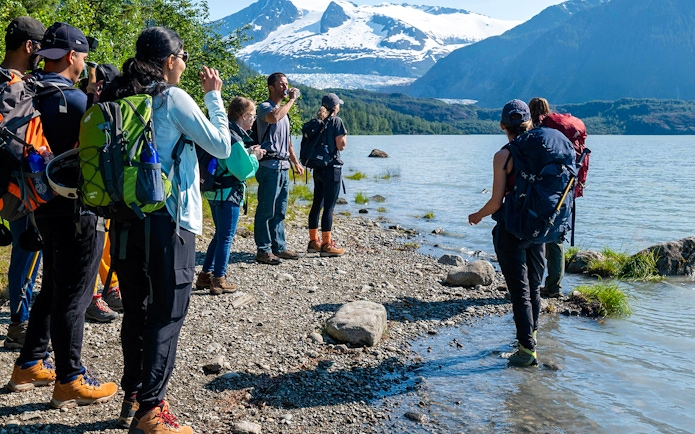 Group hiking along Mendenhall Glacier lake with snowy mountains in the background.