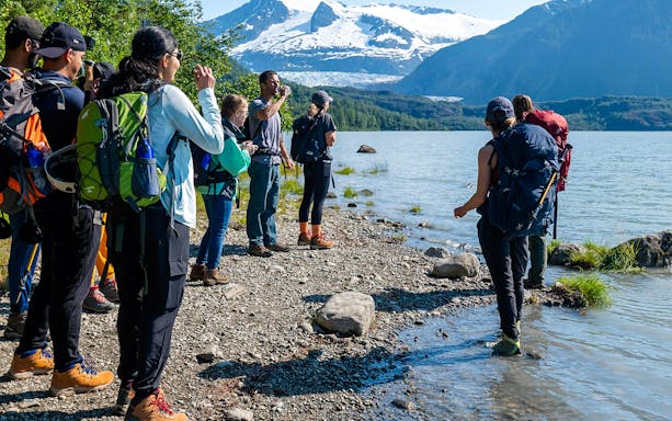 Group hiking along Mendenhall Glacier lake with snowy mountains in the background.