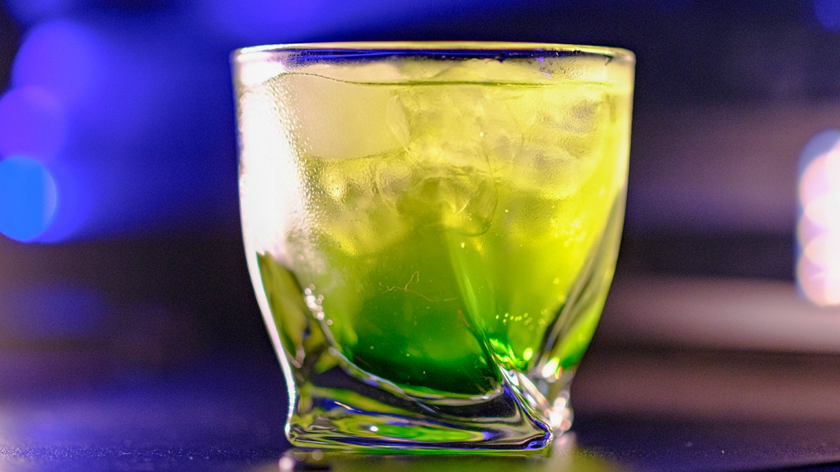 Green cocktail in a glass with ice, set against a blurred blue background.