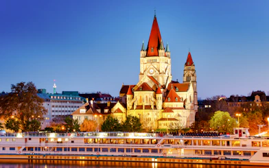 Cruise ship on Danube River with St. Francis of Assisi Church in Vienna at dusk.