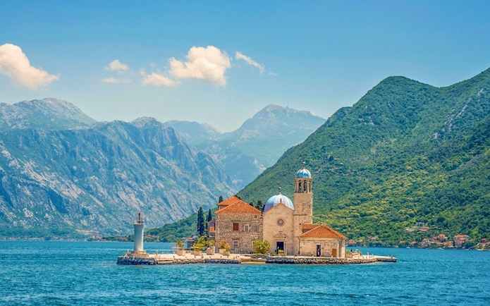 Our Lady of the Rocks island with church and lighthouse in Montenegro, surrounded by mountains.