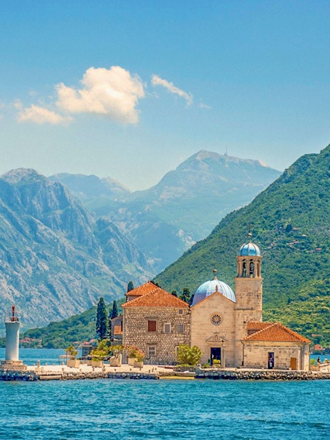 Our Lady of the Rocks island with church and lighthouse in Montenegro, surrounded by mountains.