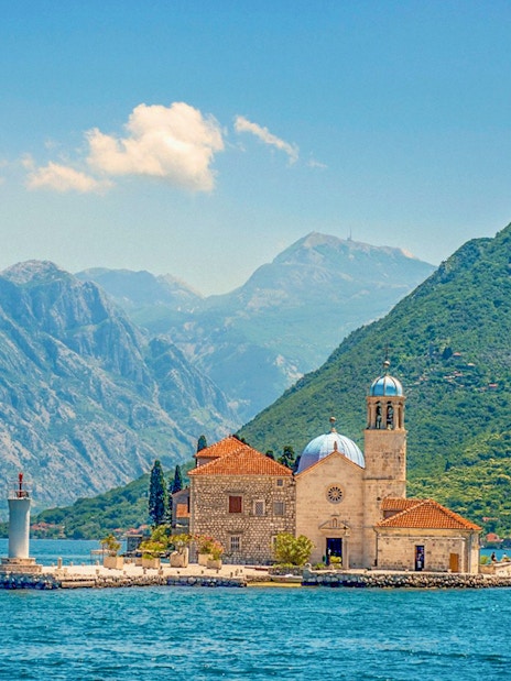 Our Lady of the Rocks island with church and lighthouse in Montenegro, surrounded by mountains.