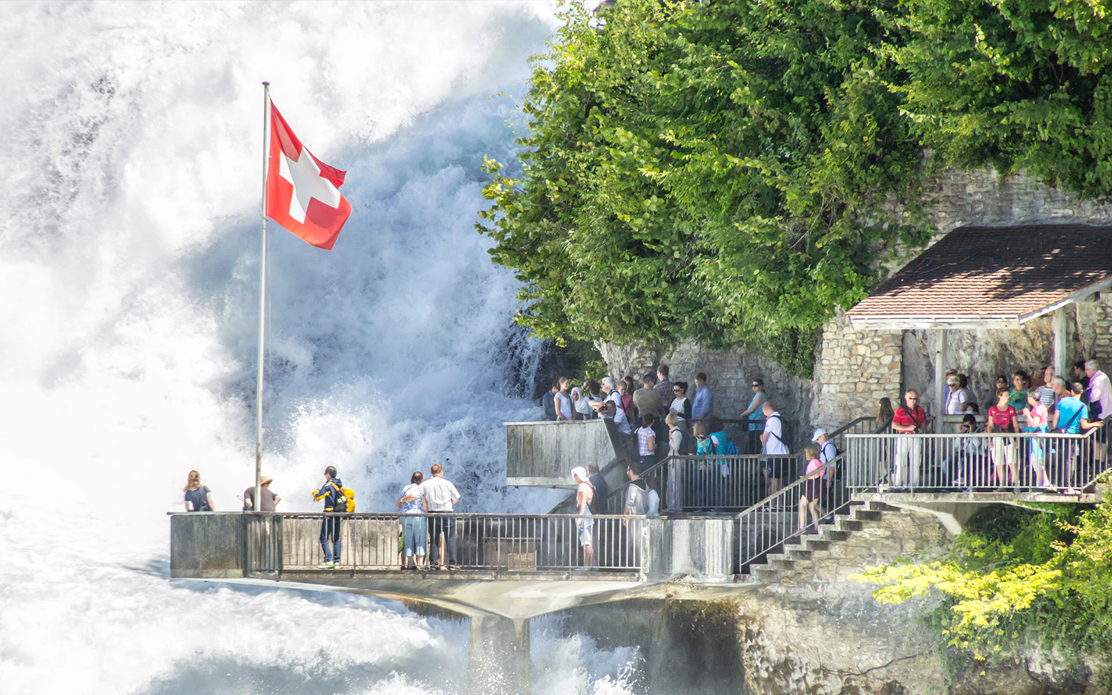 Visitors on Rhine Falls viewing platform with Swiss flag, surrounded by cascading water and lush greenery.