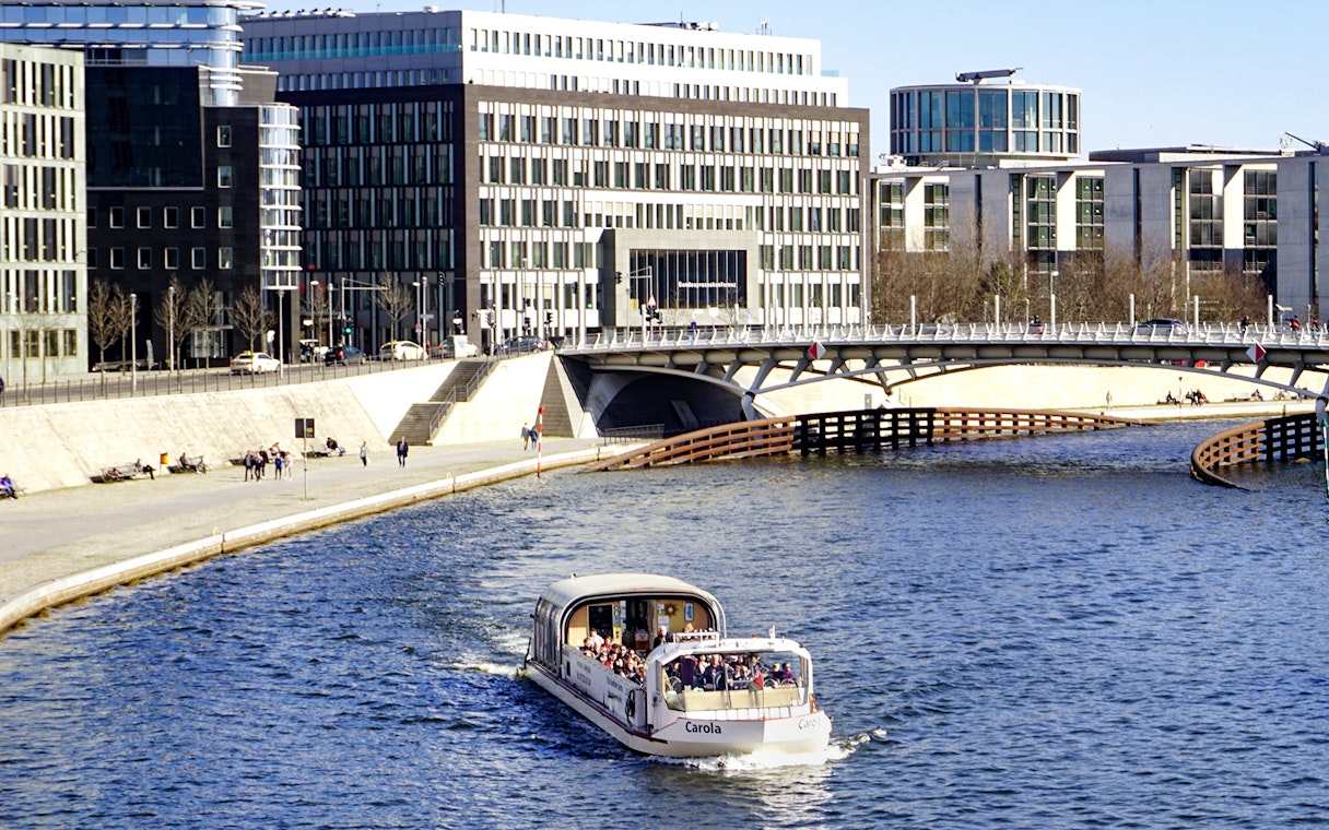 River cruise boat with tour guide on urban waterway near modern buildings.