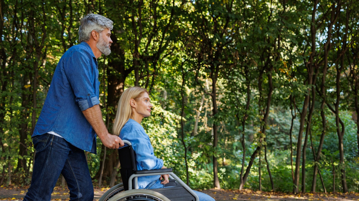 Man pushing woman in wheelchair along tree-lined path at Jardin Secret.