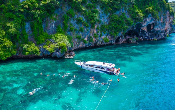 Snorkelers near a boat in clear waters by the cliffs of Phi Phi Islands.