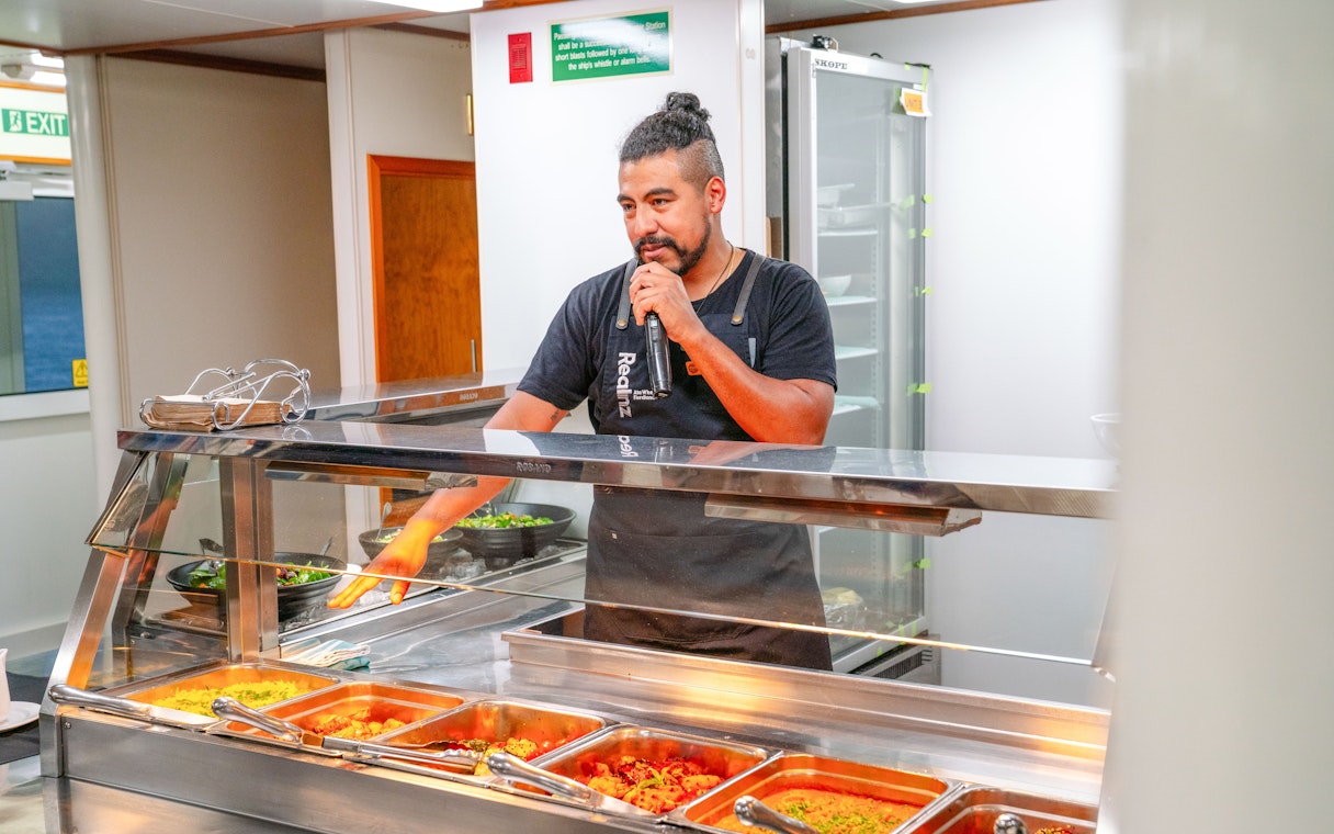 Crew member speaking to guests over a microphone behind a buffet of hot food on a boat.