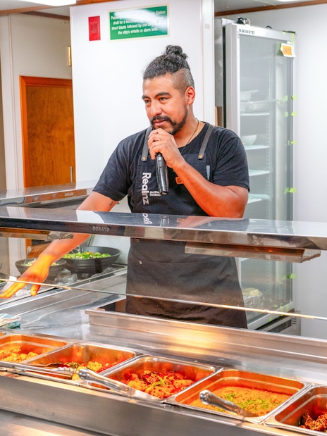Crew member speaking to guests over a microphone behind a buffet of hot food on a boat.