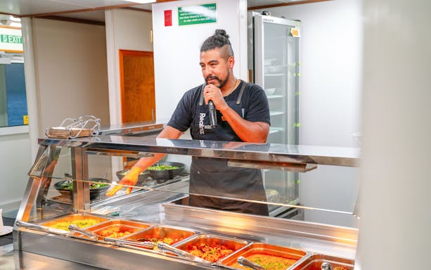 Crew member speaking to guests over a microphone behind a buffet of hot food on a boat.