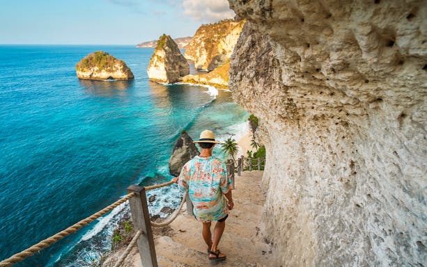 Tourist descending steps with view of Atuh Cliff and ocean on Nusa Penida Island.