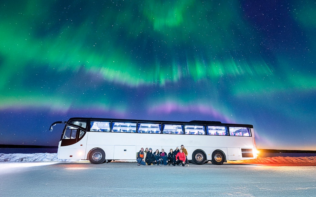 Tourists gather near a bus under the Northern Lights in a snowy landscape.