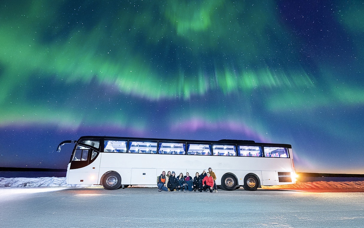 Tourists gather near a bus under the Northern Lights in a snowy landscape.