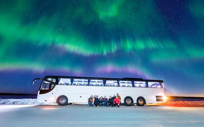 Tourists gather near a bus under the Northern Lights in a snowy landscape.