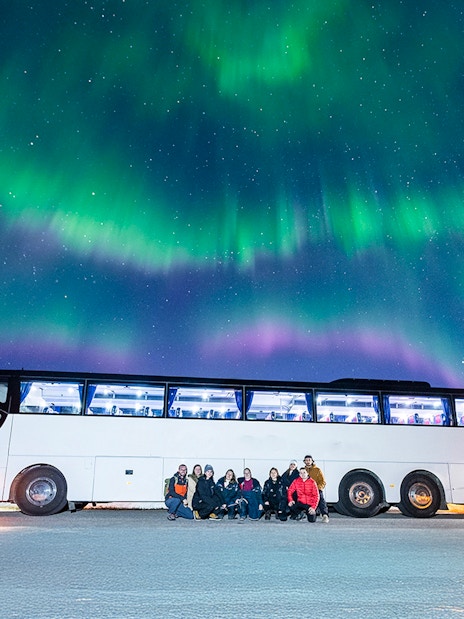 Tourists gather near a bus under the Northern Lights in a snowy landscape.