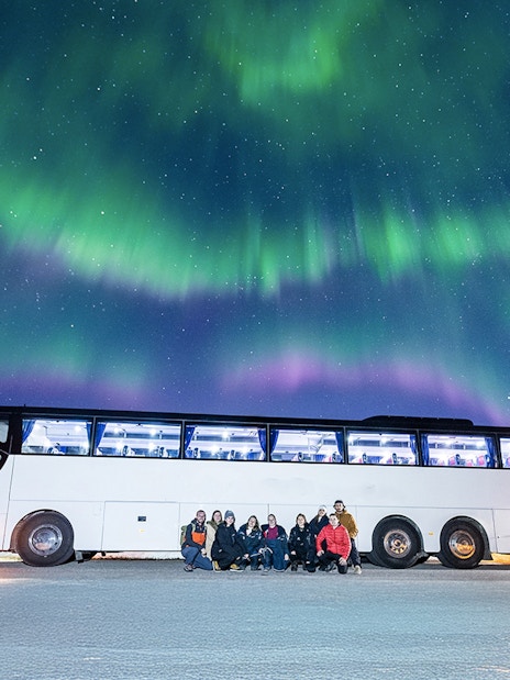 Tourists gather near a bus under the Northern Lights in a snowy landscape.