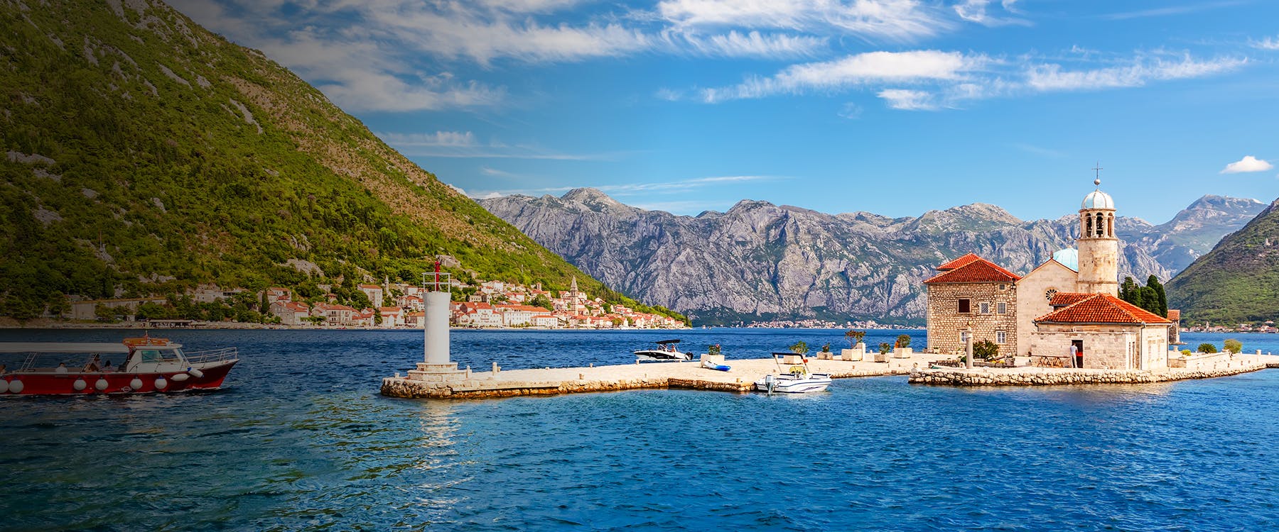 Island church and lighthouse in Tivat, Montenegro, with mountains and boats in the background.