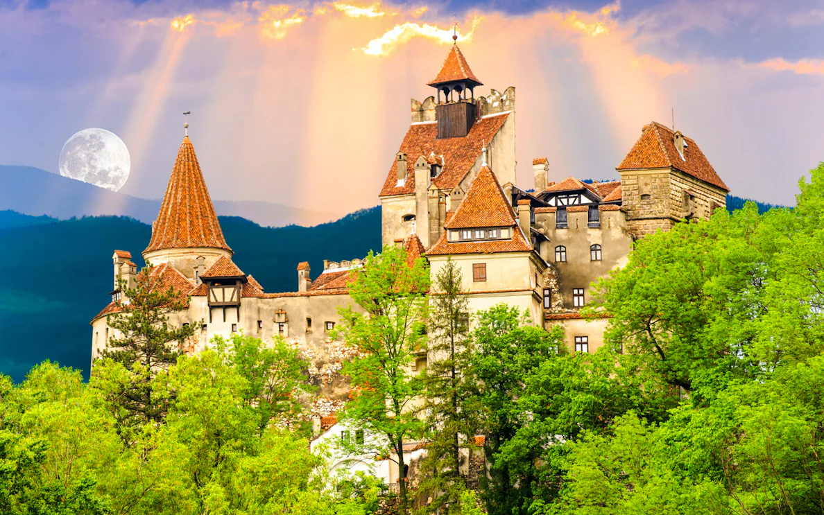 Brasov's Bran Castle surrounded by lush greenery under a full moon.