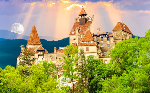 Brasov's Bran Castle surrounded by lush greenery under a full moon.