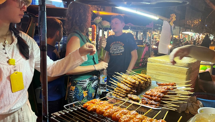 Tuk-tuk tour group enjoying street food skewers in Bangkok's Chinatown.