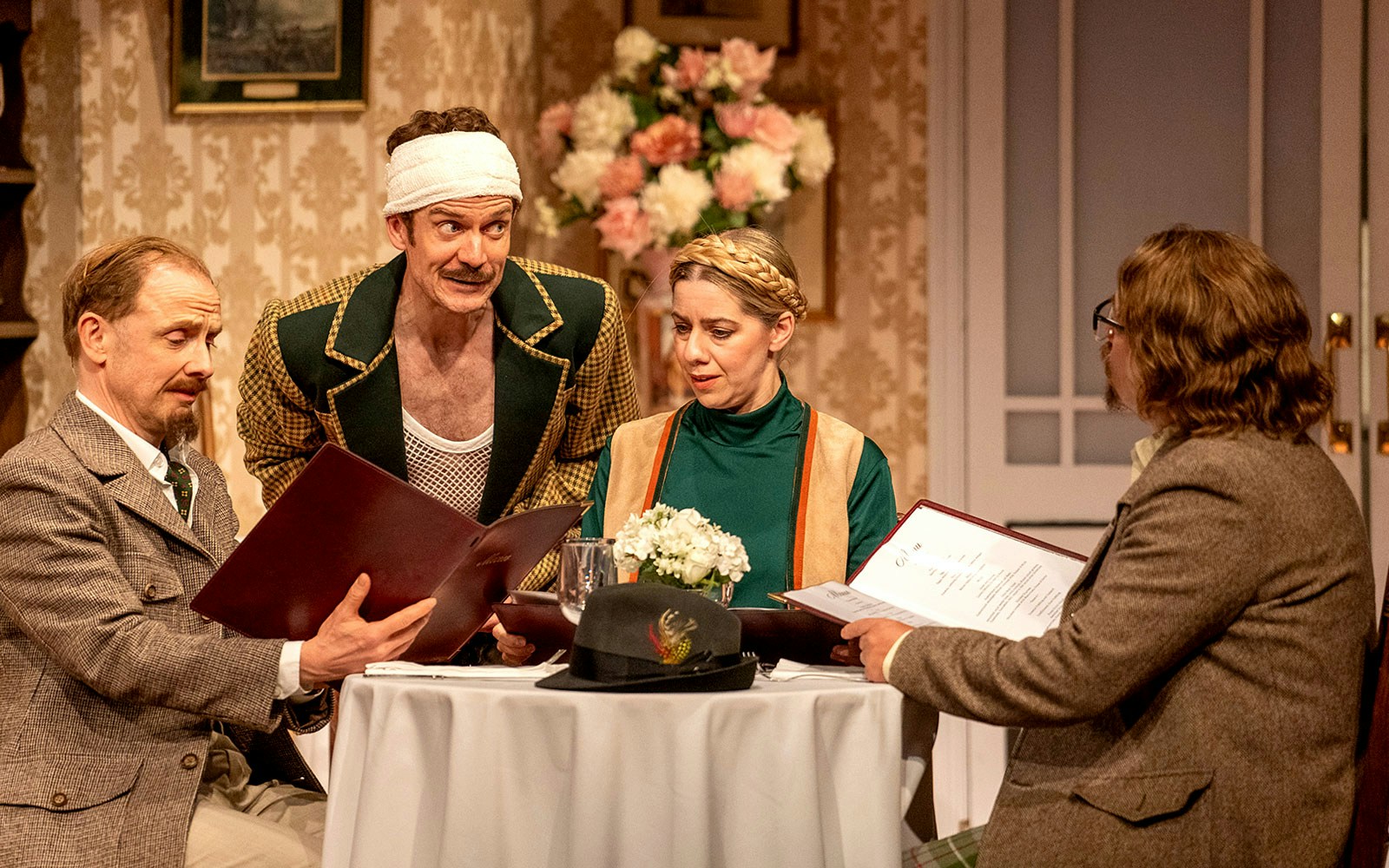 Four actors in a Fawlty Towers stage scene, seated at a table with menus.