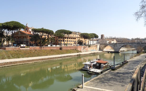 Hop-on hop-off river cruise boat docked on the Tiber River in Rome, near historic buildings and a stone bridge.