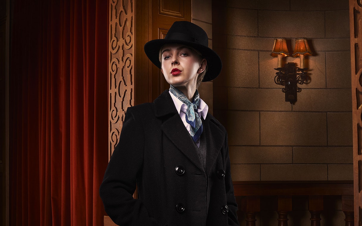 Person in vintage attire with hat and scarf, standing in a theater setting for The Mousetrap.