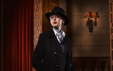 Person in vintage attire with hat and scarf, standing in a theater setting for The Mousetrap.