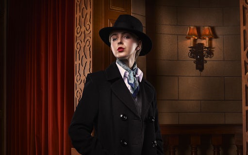 Person in vintage attire with hat and scarf, standing in a theater setting for The Mousetrap.