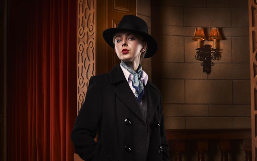 Person in vintage attire with hat and scarf, standing in a theater setting for The Mousetrap.