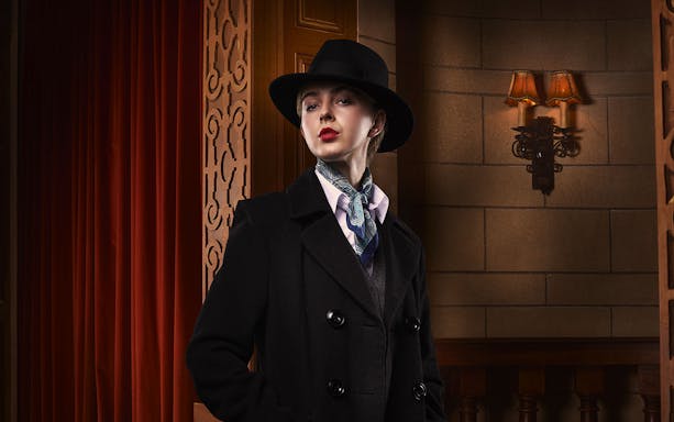 Person in vintage attire with hat and scarf, standing in a theater setting for The Mousetrap.
