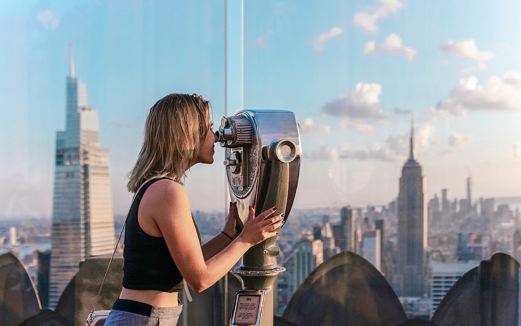 Woman using binoculars at Top of the Rock Observation Deck with Empire State Building view.