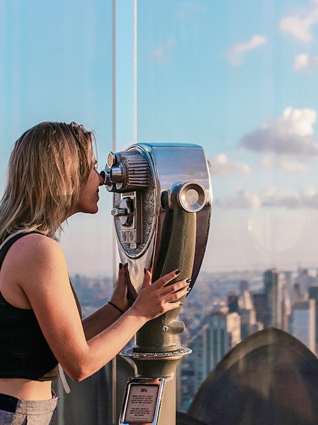 Woman using binoculars at Top of the Rock Observation Deck with Empire State Building view.