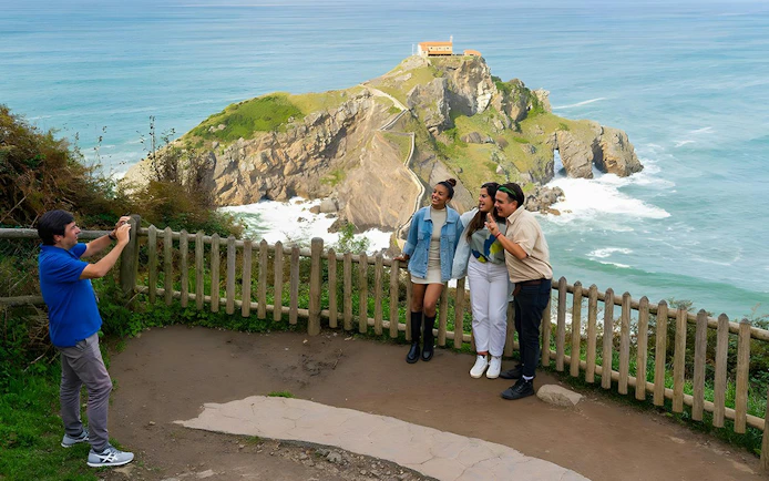 Visitors posing for a photo with San Juan de Gaztelugatxe and its stone footbridge in the background.
