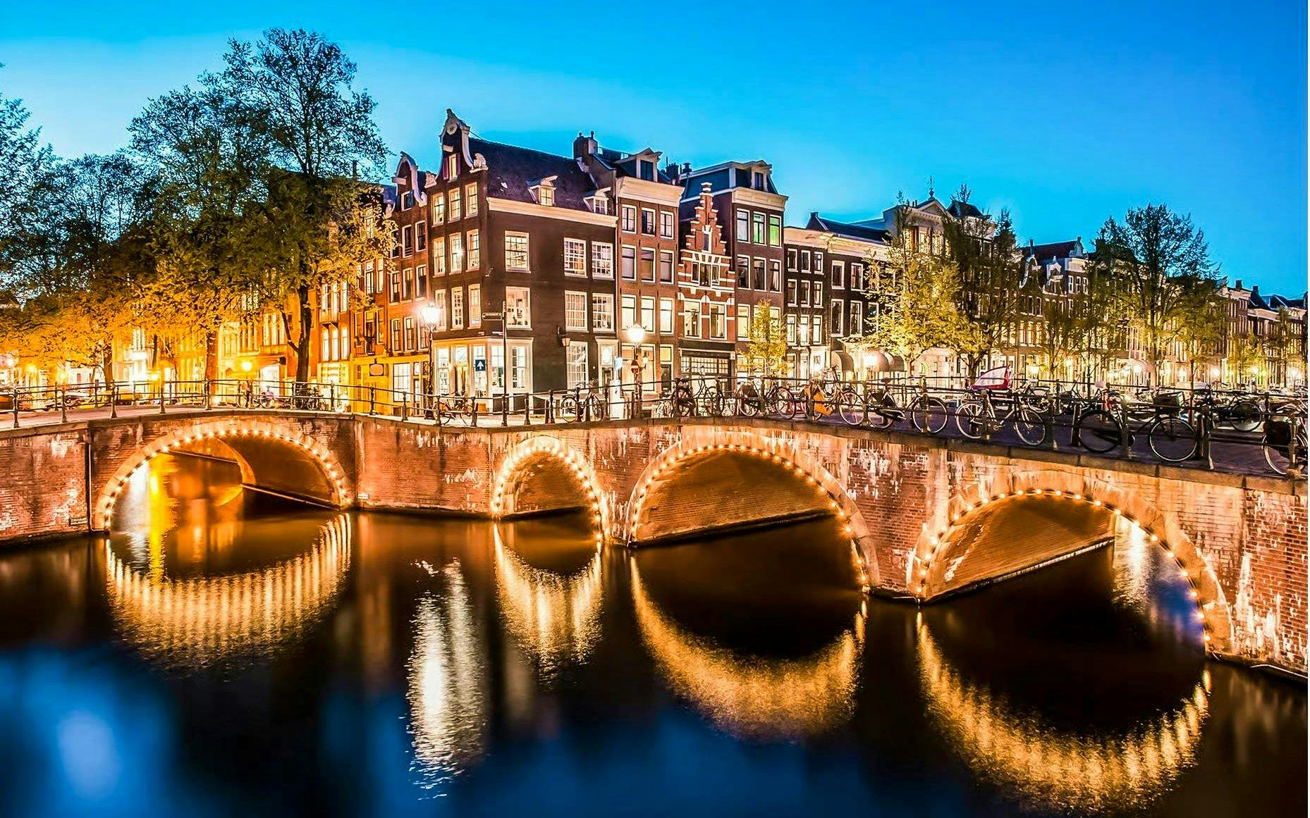 Amsterdam canal with illuminated bridge and historic buildings at dusk.