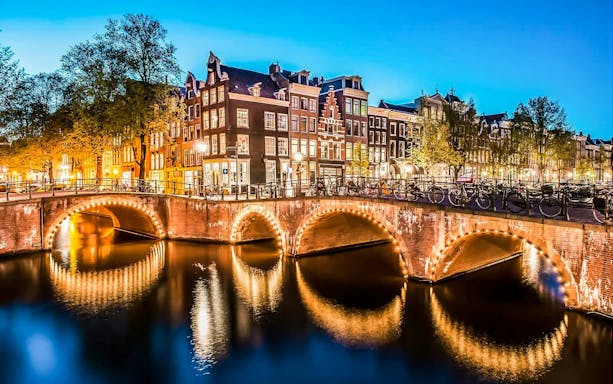 Amsterdam canal with illuminated bridge and historic buildings at dusk.