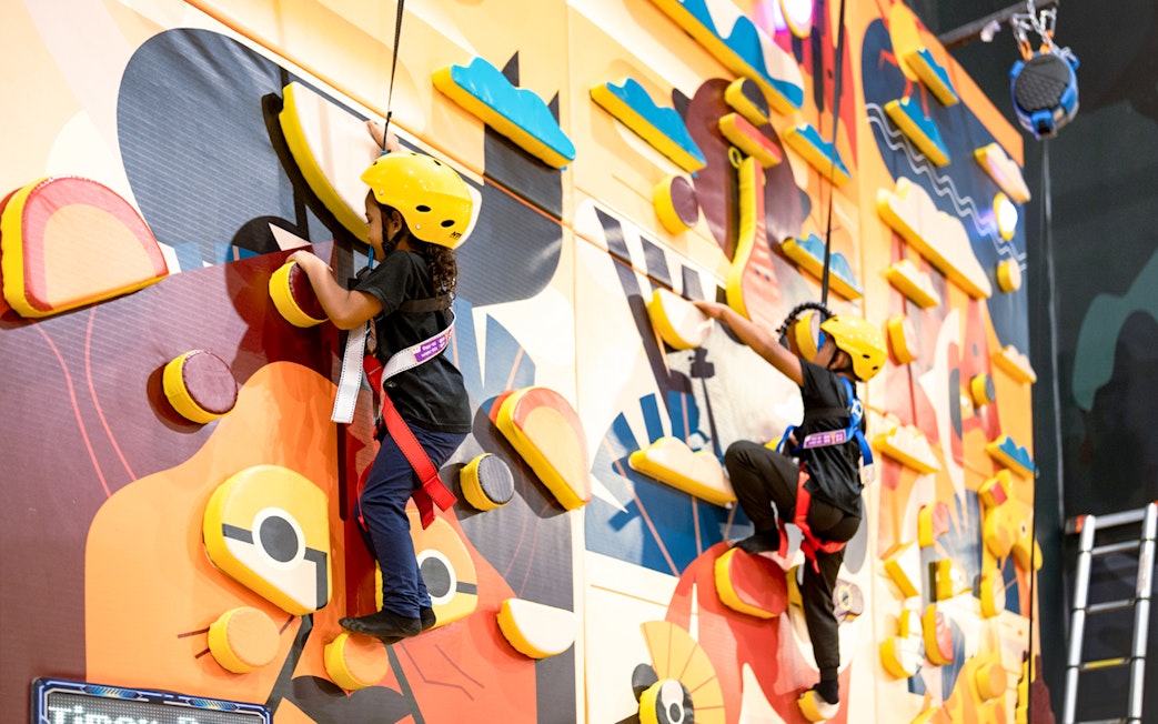 Children climbing a colorful indoor rock wall with safety harnesses.