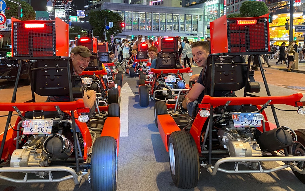 Go-kart riders on Shibuya street during 1-hour tour, Tokyo cityscape in background.
