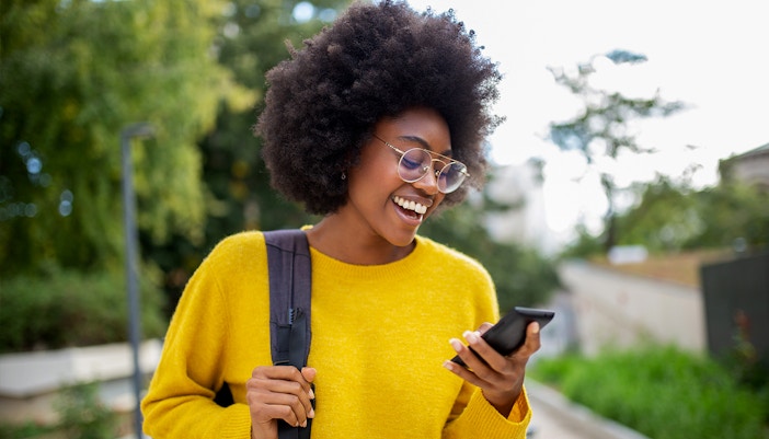 Woman with glasses smiling while checking her phone outdoors.