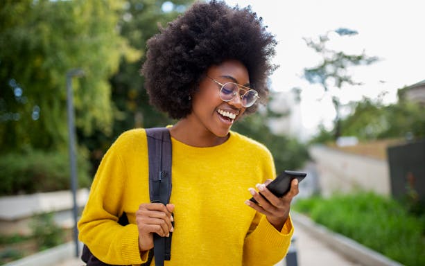 Woman with glasses smiling while checking her phone outdoors.