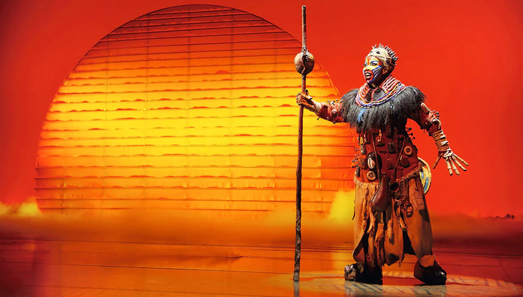 Performer in Der König der Löwen musical with sunset backdrop.