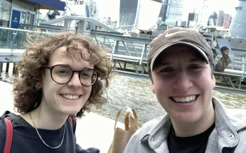 Two people smiling by the Thames River with London skyline in the background.