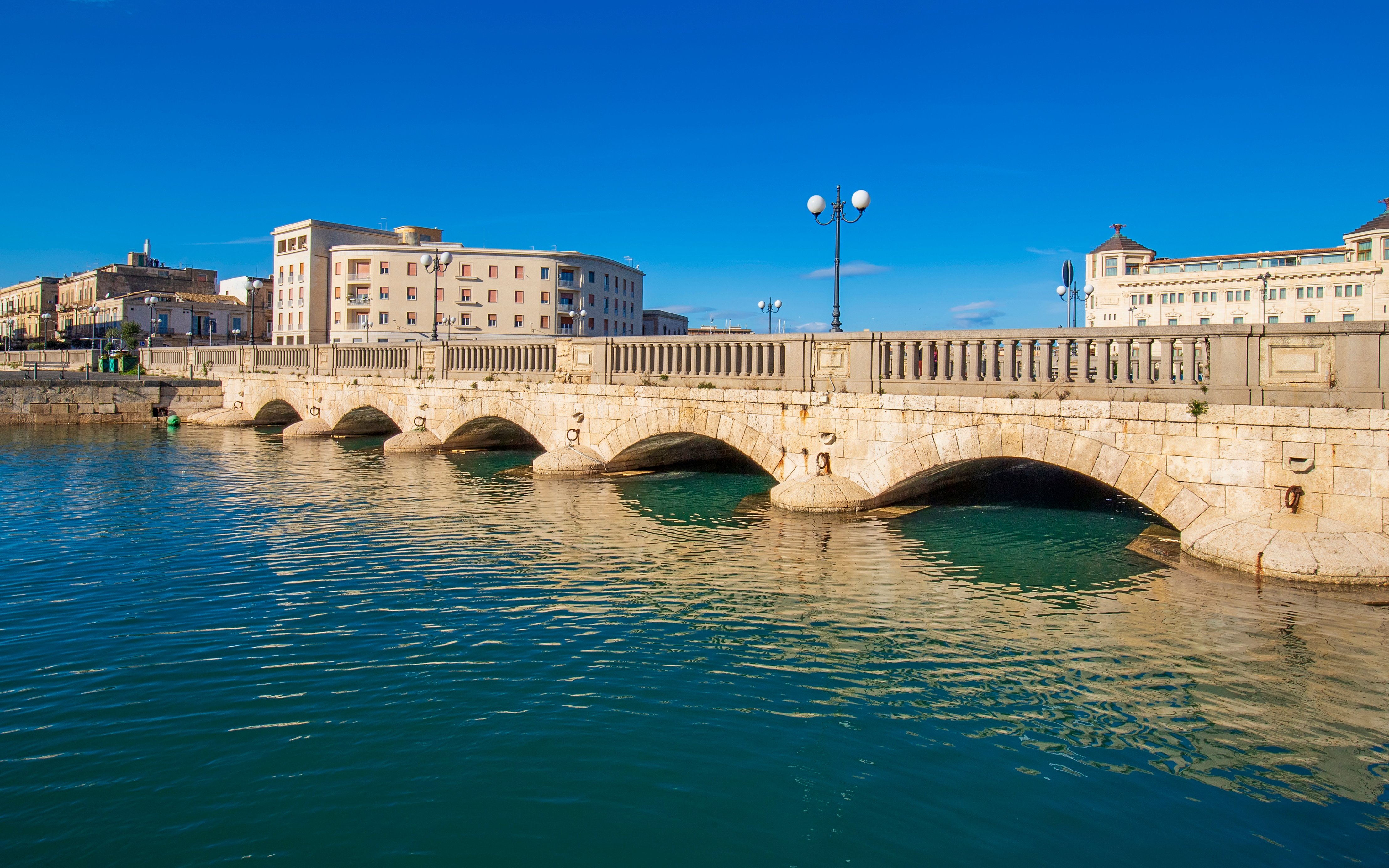 Umberto I Bridge over water in Syracuse, Sicily, Italy with buildings in the background.