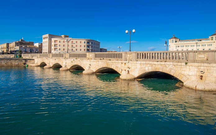 Umberto I Bridge over water in Syracuse, Sicily, Italy with buildings in the background.